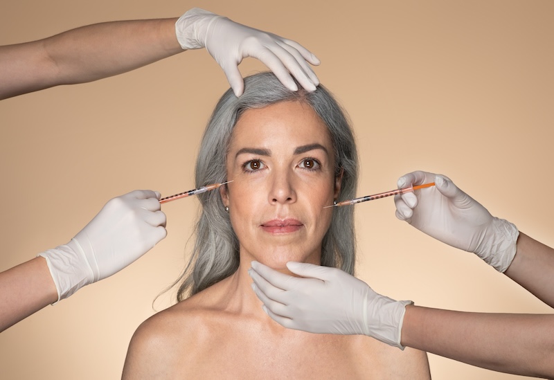 Beautiful senior woman getting facial injections by two cosmetologists, beautician hands in gloves giving anti aging serum shots on aged female face, beige studio background